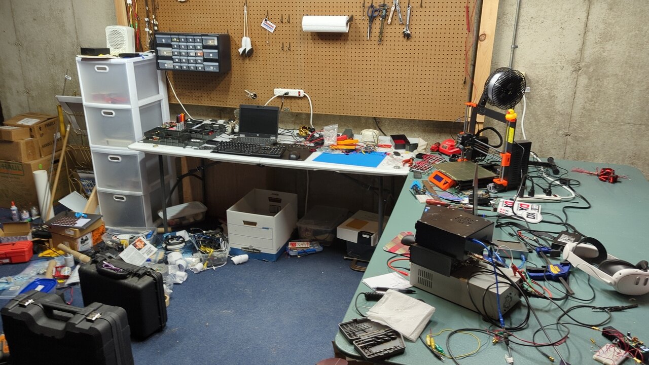 A messy lab space; tools, boxes, parts, and pieces are all over the floor and tables with almost no organization